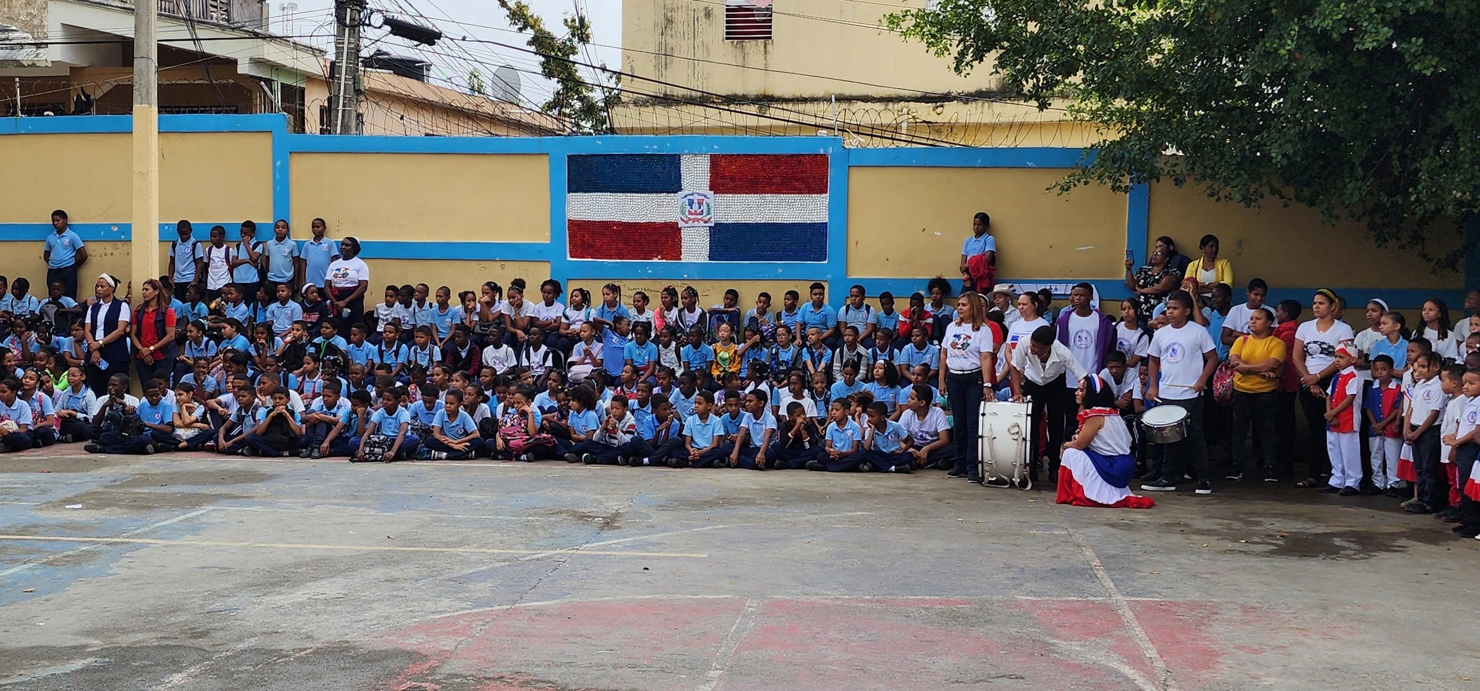 Students and staff of Escuela Primaria Jesús Maestro gathered in the schoolyard, with the Dominican Republic flag painted on the wall behind them.